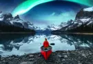a single person sitting in a canoe at the edge of a lake with mountains in the distance and northern lights in the sky