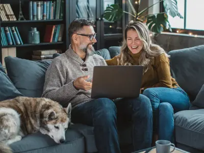 Man and woman sitting on a couch with their dog sipping coffee and looking at tablet