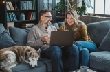 Man and woman sitting on a couch with their dog sipping coffee and looking at tablet