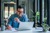 Man smiling and working at his laptop and writing notes beside